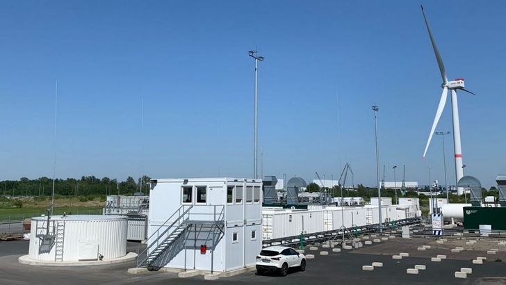 A renewable energy facility with white buildings, a wind turbine, and a parked white car under a clear blue sky. A renewable energy facility with white buildings, a wind turbine, and a parked white car under a clear blue sky.
