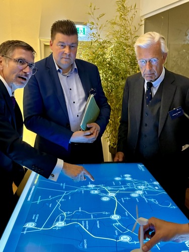 Three men in suits discuss a digital map displayed on a large touchscreen table.