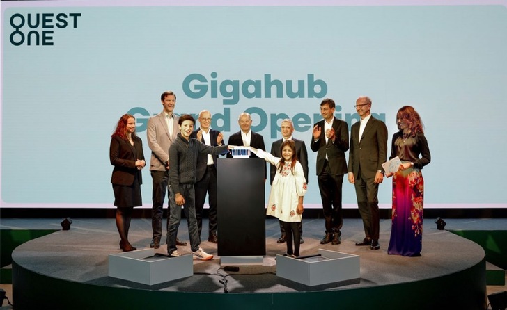 Ten people stand on stage at the Gigahub Grand Opening, with two children pressing a button at the center. Ten people stand on stage at the Gigahub Grand Opening, with two children pressing a button at the center.