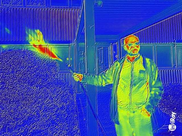 A man in a heat map effect image uses a torch to burn a bush outside a building.