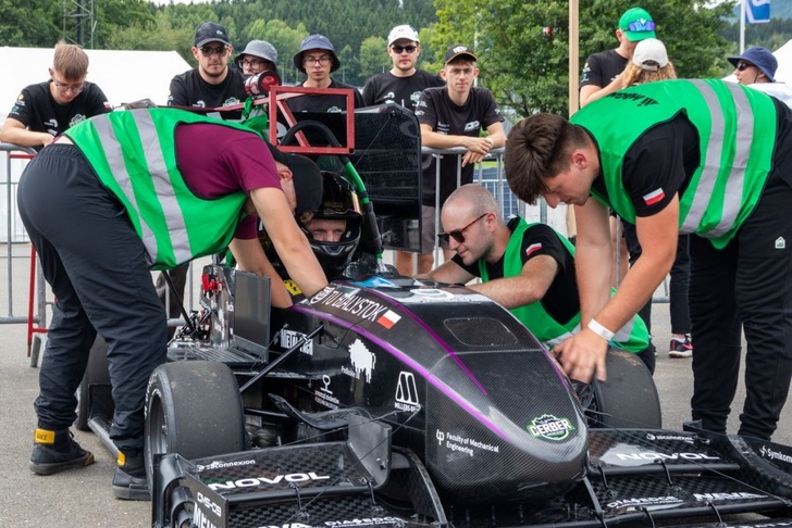 Three crew members in green vests work on a race car as a team of people watch in the background. Three crew members in green vests work on a race car as a team of people watch in the background.