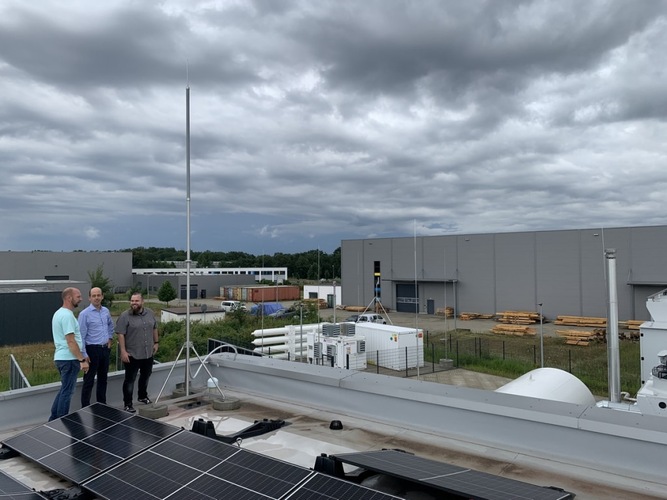 Three men stand on a rooftop with solar panels, industrial buildings and cloudy sky in the background. Three men stand on a rooftop with solar panels, industrial buildings and cloudy sky in the background.