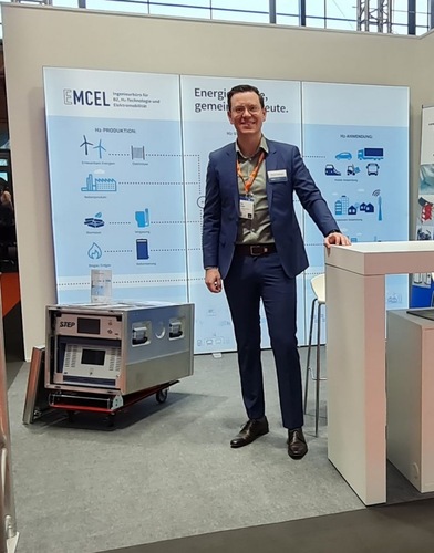 A man in a suit stands at an exhibition booth with technical equipment and informational displays behind him. A man in a suit stands at an exhibition booth with technical equipment and informational displays behind him.