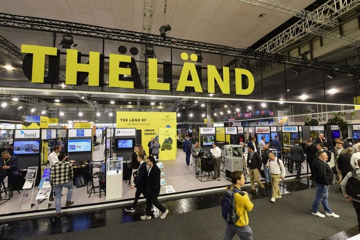 People walk through a busy tech expo with a large “THE LÄND” sign overhead and display booths below. People walk through a busy tech expo with a large “THE LÄND” sign overhead and display booths below.