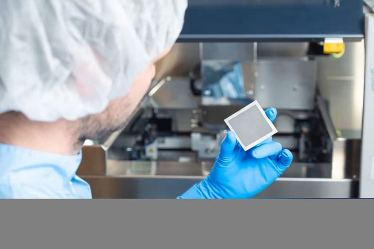 A scientist in gloves and a hairnet holds a small square device in a laboratory setting. A scientist in gloves and a hairnet holds a small square device in a laboratory setting.